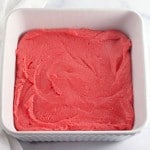 A white square baking dish filled with smooth, swirled pink batter, reminiscent of Strawberry Crunch Bars, rests on a marble surface next to a white cloth.