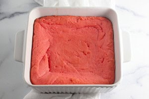 A square white baking dish cradles a delightful pink cake, reminiscent of Strawberry Crunch Bars, resting elegantly on a marble surface.