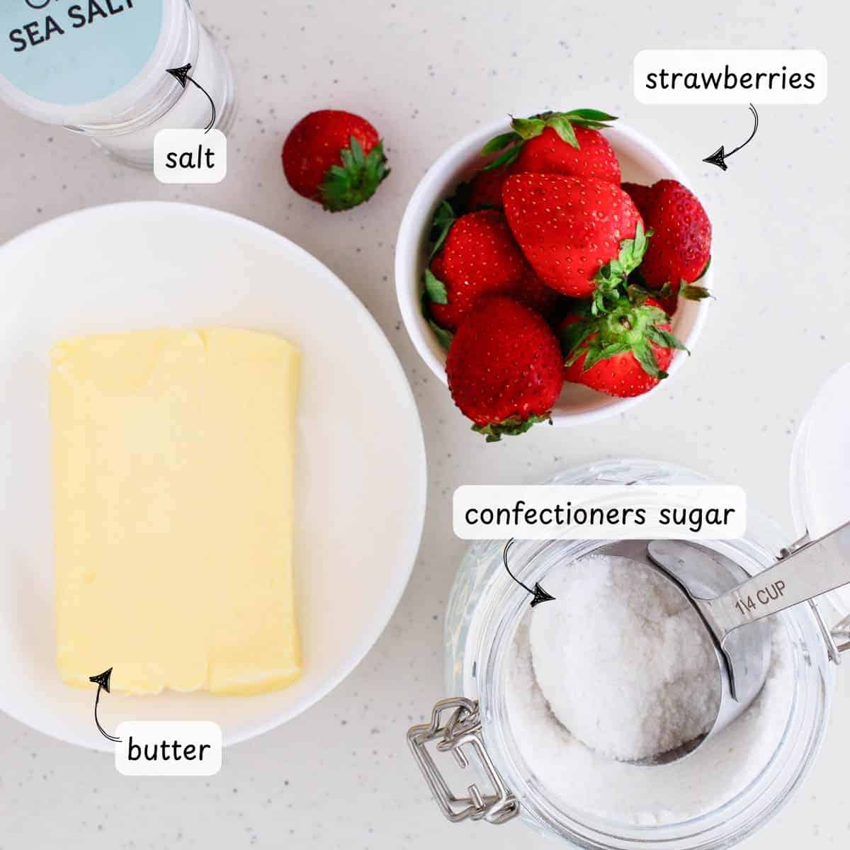 Ingredients laid out on a countertop: a plate of strawberry butter, a jar of confectioners sugar with a scoop, a bowl of strawberries, and a container of salt.