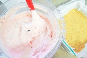 A mixing bowl with pink batter and a red spatula next to a container holding a crumbly yellow mixture.