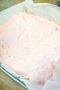A square baking tray filled with smooth, pink strawberry cheesecake mixture lined with parchment paper.
