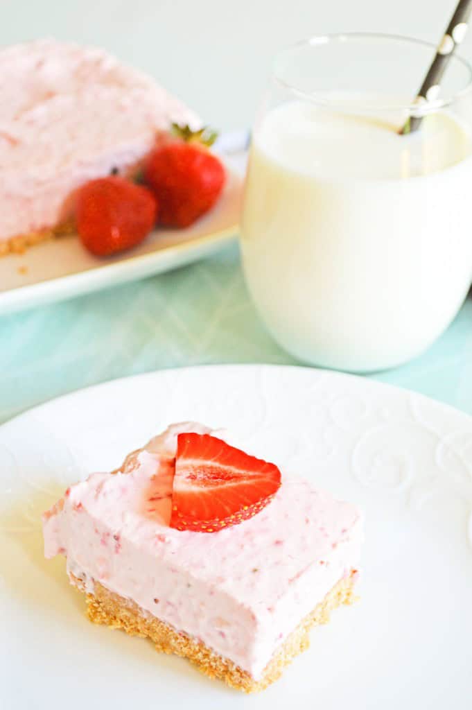 A slice of strawberry cheesecake on a white plate, topped with a strawberry piece, with whole strawberries and a glass of milk in the background.