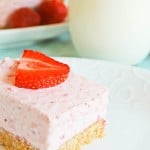 A slice of strawberry cheesecake with a graham cracker crust on a white plate, topped with a halved strawberry, accompanied by a glass of milk in the background.