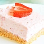 A slice of strawberry cheesecake with a crumbly base, topped with a strawberry slice, on a white plate.