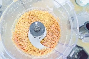 Orange mixture in a food processor bowl with a metal blade attachment, viewed from above.