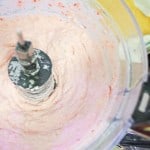 Close-up of a food processor blending pink cream mixture, with some ingredients visible in the background.