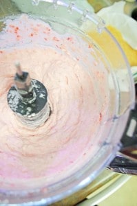 Close-up of a food processor blending pink cream mixture, with some ingredients visible in the background.