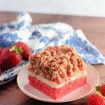 A slice of strawberry cheesecake crumble on a plate, with fresh strawberries, reminiscent of strawberry crunch bars. It boasts a red base, creamy middle layer, and crumbly top. A patterned cloth is in the background.