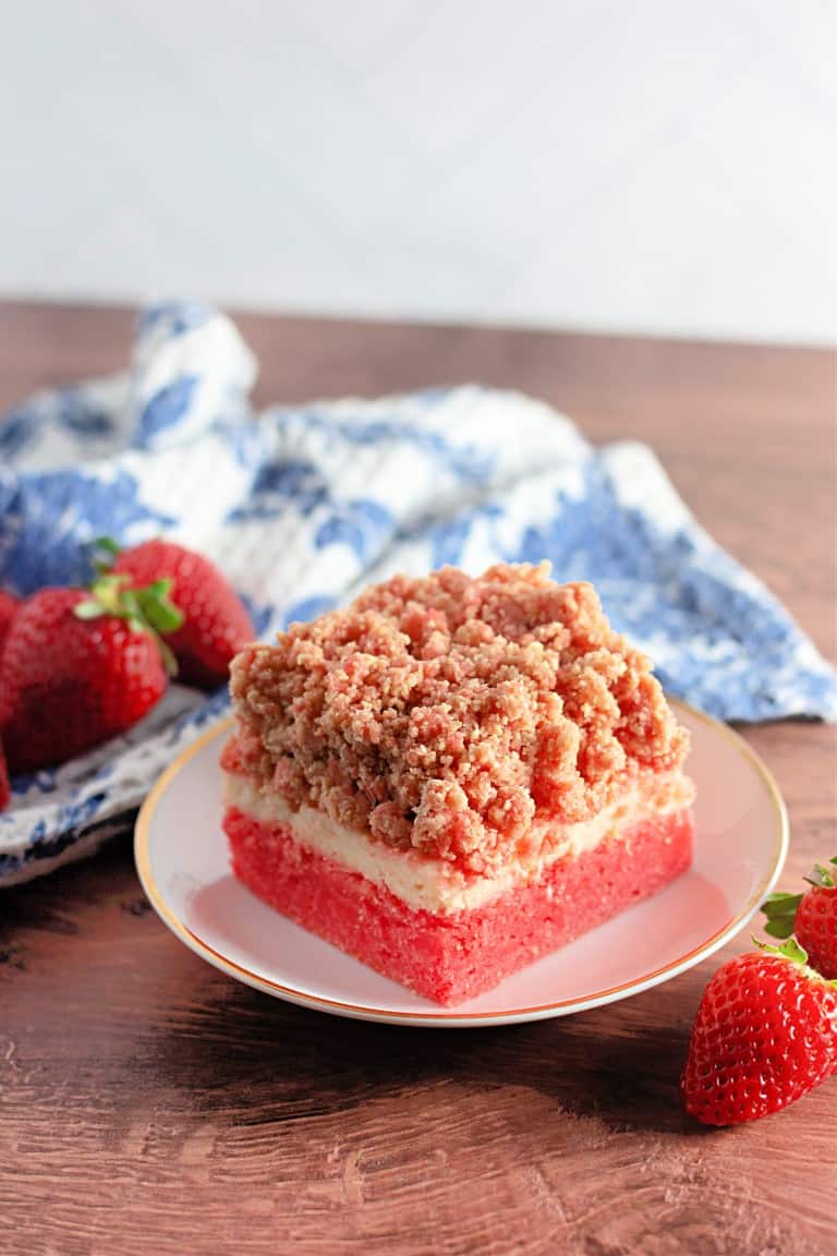 A slice of strawberry cheesecake crumble on a plate, with fresh strawberries, reminiscent of strawberry crunch bars. It boasts a red base, creamy middle layer, and crumbly top. A patterned cloth is in the background.