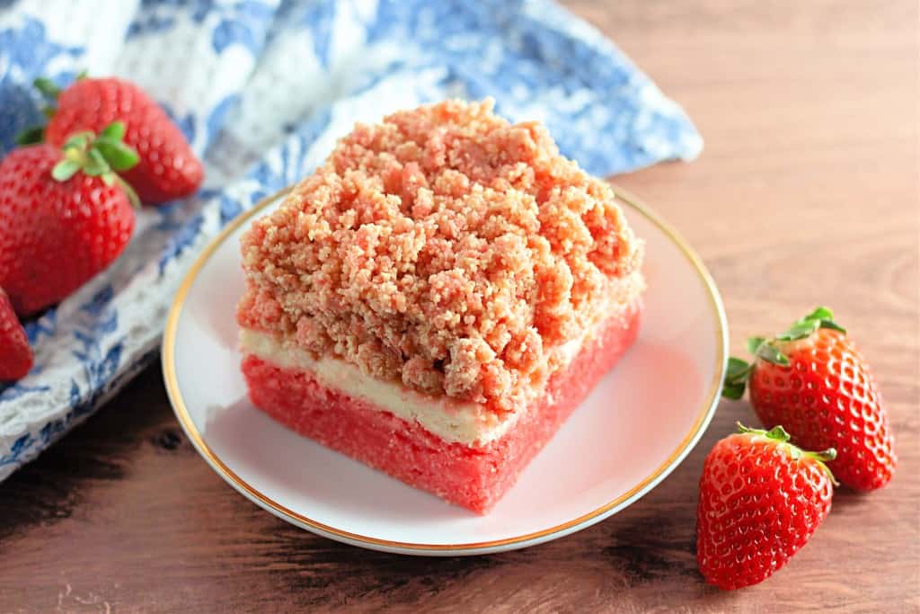 A square piece of strawberry crumb cake, reminiscent of delightful Strawberry Crunch Bars, sits on a plate, flanked by fresh strawberries and a blue-patterned cloth.