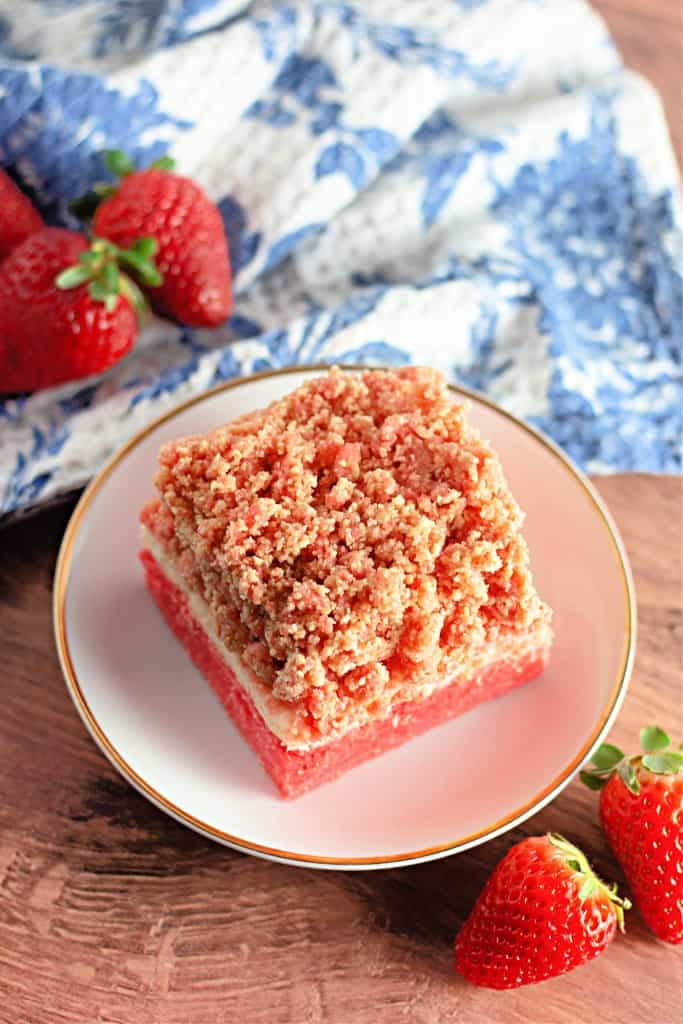 A slice of strawberry crumb cake graces a white plate, embraced by fresh strawberries and a blue-patterned cloth. It captures the essence of delightful strawberry crunch bars on a rustic wooden surface.