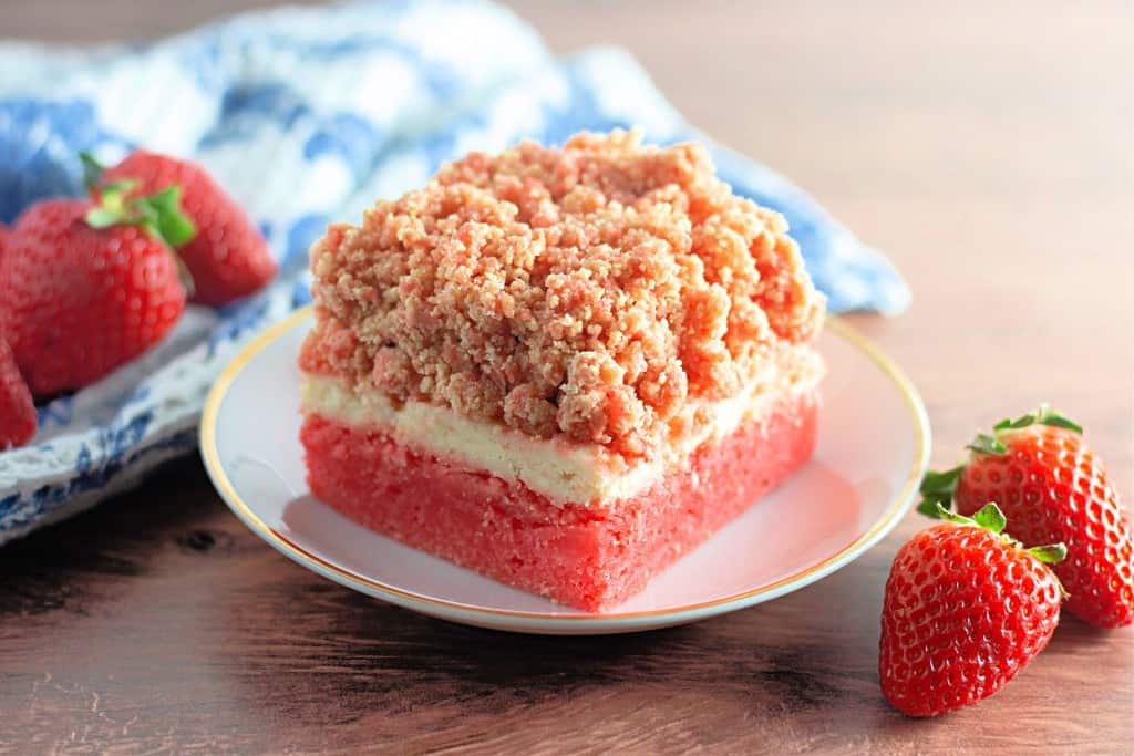 A slice of strawberry crumb cake, reminiscent of indulgent strawberry crunch bars, sits invitingly on a plate, accompanied by two fresh strawberries and a quaint blue and white cloth in the background.