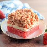 A slice of strawberry crumb cake, reminiscent of indulgent strawberry crunch bars, sits invitingly on a plate, accompanied by two fresh strawberries and a quaint blue and white cloth in the background.