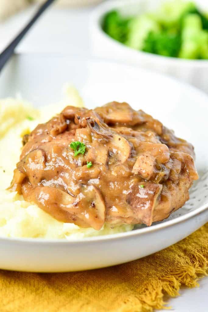 A plate featuring succulent Turkey Salisbury Steak nestled beside mashed potatoes topped with mushroom gravy and herbs, with vibrant broccoli in the background.