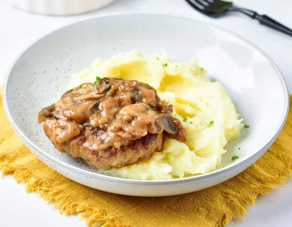 A plate with mashed potatoes topped with a turkey Salisbury steak and mushroom gravy, placed on a yellow cloth napkin. A fork is in the background.