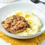 A plate with mashed potatoes topped with a turkey Salisbury steak and mushroom gravy, placed on a yellow cloth napkin. A fork is in the background.