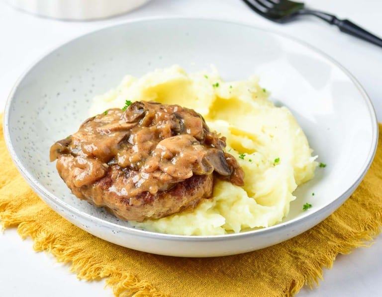 A plate with mashed potatoes topped with a turkey Salisbury steak and mushroom gravy, placed on a yellow cloth napkin. A fork is in the background.