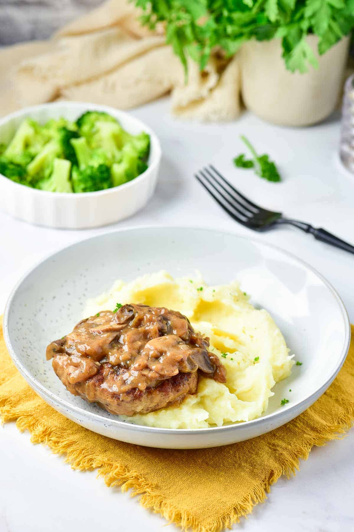 A plate with mashed potatoes topped with a turkey Salisbury steak and mushroom gravy, placed on a yellow cloth napkin. A fork is in the background.