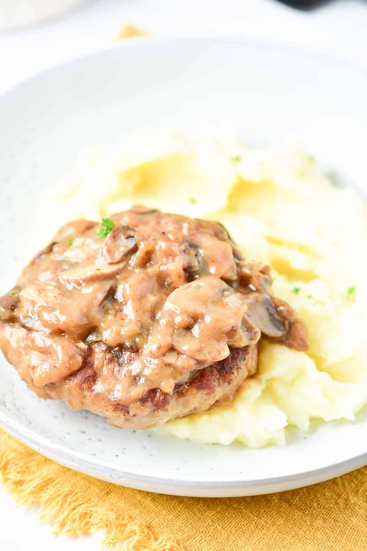 A plate with mashed potatoes topped with a turkey Salisbury steak and mushroom gravy, placed on a yellow cloth napkin. A fork is in the background.