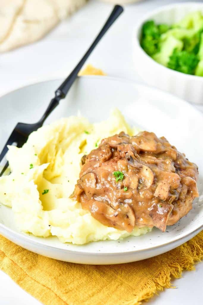 A plate with mashed potatoes topped with a savory brown mushroom and turkey Salisbury steak sauce, accompanied by a fork, and a bowl of broccoli in the background.