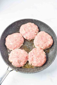 Five raw turkey burger patties sizzle in a black frying pan on the stovetop, reminiscent of a classic Salisbury steak preparation.