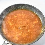 A non-stick frying pan on a stove holds a simmering brown liquid with small visible chunks, resembling the rich gravy of a Turkey Salisbury Steak in the process of cooking.
