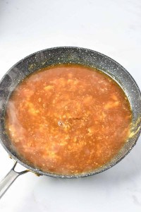 A non-stick frying pan on a stove holds a simmering brown liquid with small visible chunks, resembling the rich gravy of a Turkey Salisbury Steak in the process of cooking.