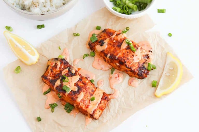 Two grilled salmon fillets topped with a creamy sauce and chopped green onions rest elegantly on parchment paper, garnished with lemon wedges—a perfect addition to your collection of seafood recipes.