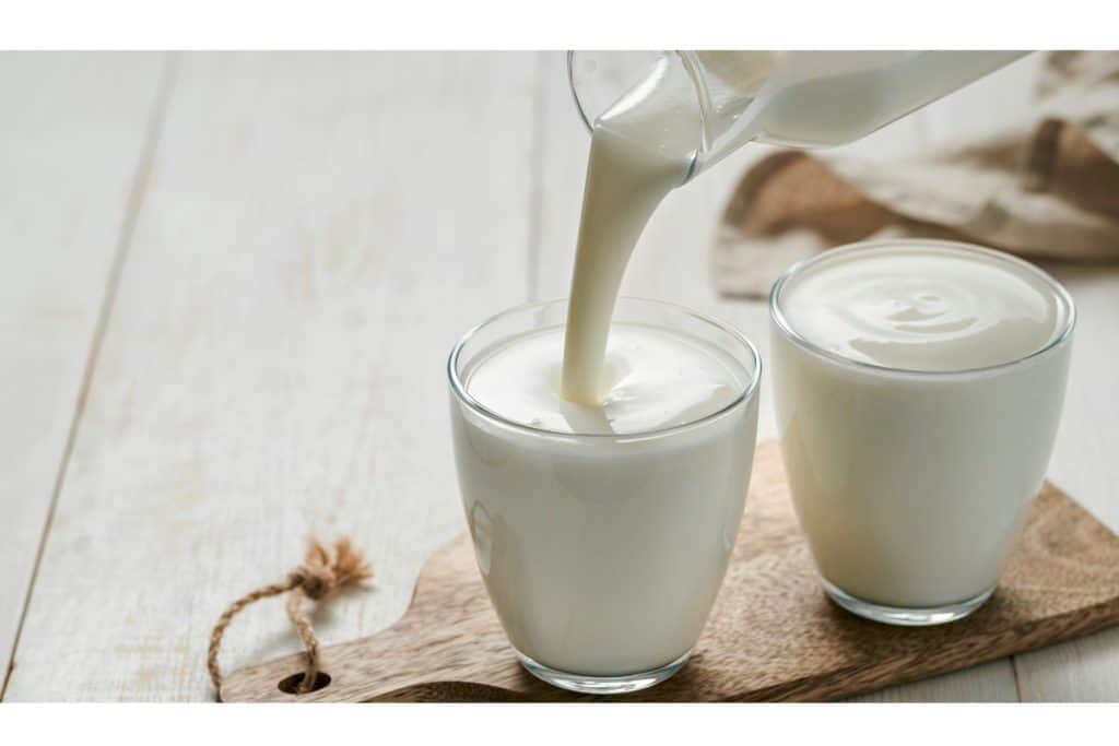 Milk being poured into a glass on a wooden board, next to another filled glass, on a light-colored surface.