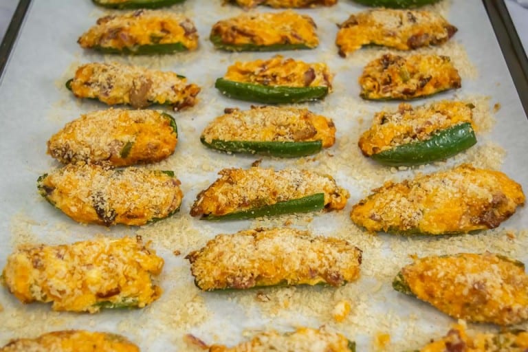 Twenty baked jalapeño poppers with melted cheese and breadcrumbs on a parchment-lined baking sheet.