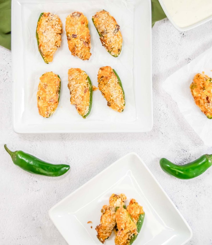 Three plates with stuffed jalapeños, two whole jalapeños, and a green napkin on a white surface.