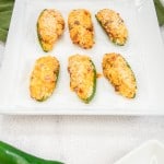 Six baked jalapeño poppers topped with breadcrumbs on a white square plate, with a green napkin in the background and one whole jalapeño pepper in the foreground.