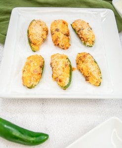 Six baked jalapeño poppers topped with breadcrumbs on a white square plate, with a green napkin in the background and one whole jalapeño pepper in the foreground.