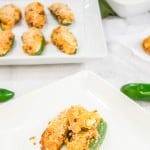 Two plated jalapeño poppers on a white dish with more poppers on a tray in the background.