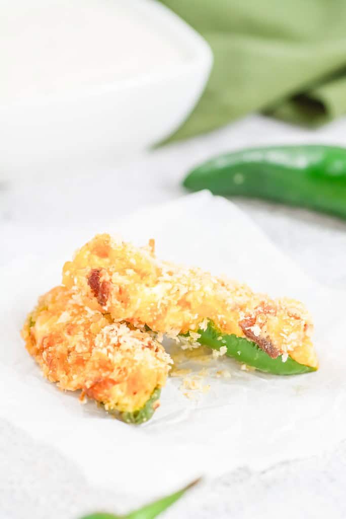 Two breaded jalapeño poppers on parchment paper, with a green jalapeño and white sauce dish in the background.
