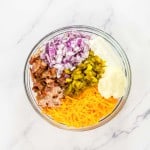 Bowl with chopped red onions, bacon, pickles, mayonnaise, sour cream, and shredded cheddar cheese on a marble countertop.