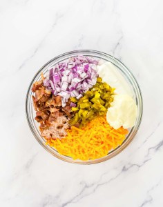 Bowl with chopped red onions, bacon, pickles, mayonnaise, sour cream, and shredded cheddar cheese on a marble countertop.