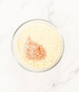 Bowl of grated Parmesan cheese with a small pile of paprika on top, placed on a white marble surface.