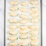 A baking sheet with 24 breaded zucchini halves topped with a crumbly mixture, arranged in rows on parchment paper.