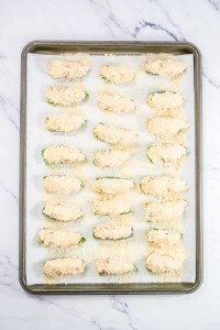 A baking sheet with 24 breaded zucchini halves topped with a crumbly mixture, arranged in rows on parchment paper.