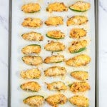 A baking sheet with 25 stuffed jalapeño halves topped with breadcrumbs on parchment paper, placed on a marble countertop.