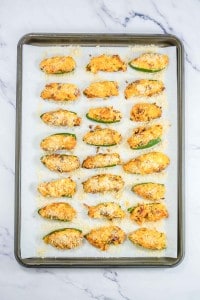 A baking sheet with 25 stuffed jalapeño halves topped with breadcrumbs on parchment paper, placed on a marble countertop.