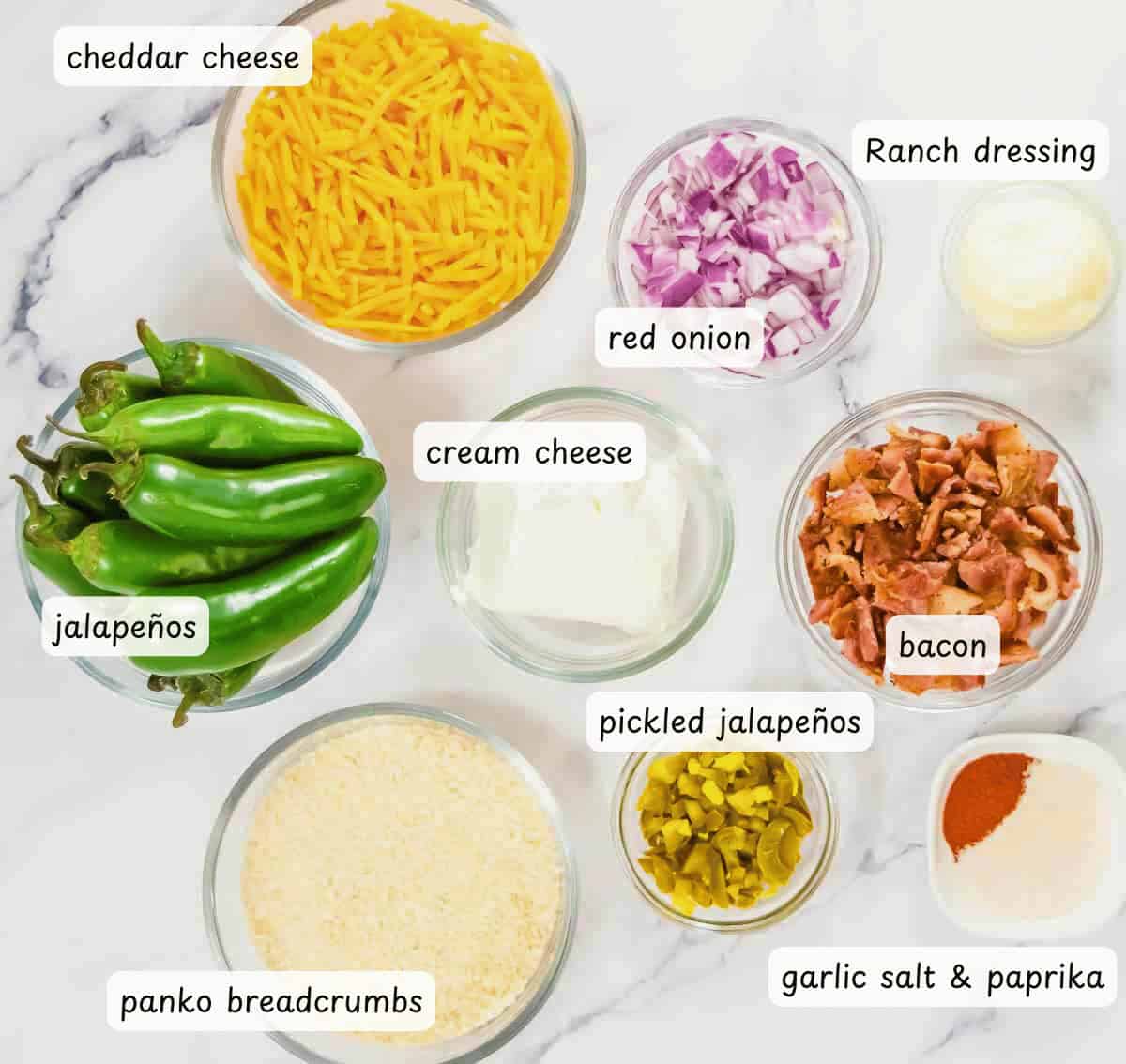 Ingredients for Air Fryer Jalapeño Poppers arranged on a marble surface include cheddar cheese, onions, fresh and pickled jalapeños, ranch dressing, cream cheese, crisp bacon, breadcrumbs, and spices.