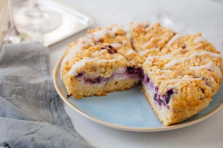 A sliced blueberry coffee cake with crumbly topping and icing drizzles sits on a plate next to a gray napkin.