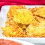 Close-up of breaded and fried food pieces on a white plate, with a bowl of red sauce in the foreground. A red cloth is visible in the background.
