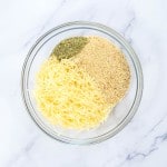 A glass bowl contains shredded cheese, dried herbs, and breadcrumbs on a white marble surface.