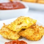 Breaded, fried ravioli on a white plate with marinara sauce, one ravioli is cut open to show filling.