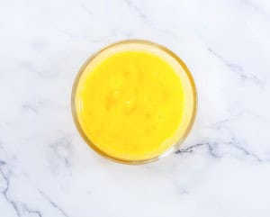 A glass bowl filled with beaten yellow egg yolks, placed on a white marble surface.