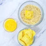 Ravioli pieces on a marble surface with three bowls: beaten eggs, breading mixture, and shredded cheese.