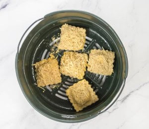 Five breaded square pieces topped with cheese are arranged in a circular way inside a dark green air fryer basket on a white marble surface.
